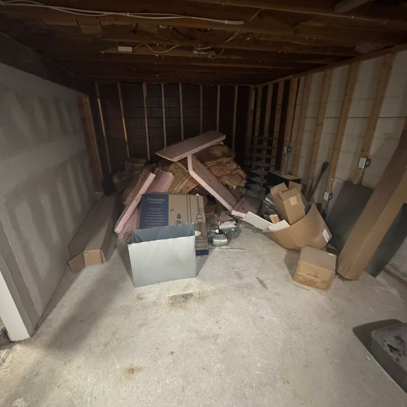 An unfinished basement corner with open framing, leaning drywall and a pile of insulation scraps and boxes