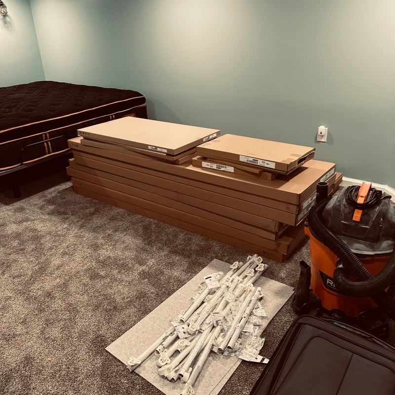 A carpeted bedroom with a bed, boxed panels on the floor and a shop vacuum